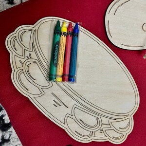 Puede incluir: Un tablero de manualidades de madera con forma de pandereta con un juego de cuatro crayones en verde, amarillo, rojo y azul envueltos en pl&aacute;stico transparente. Los crayones est&aacute;n etiquetados como "Crayons". El tablero es de madera sin terminar.