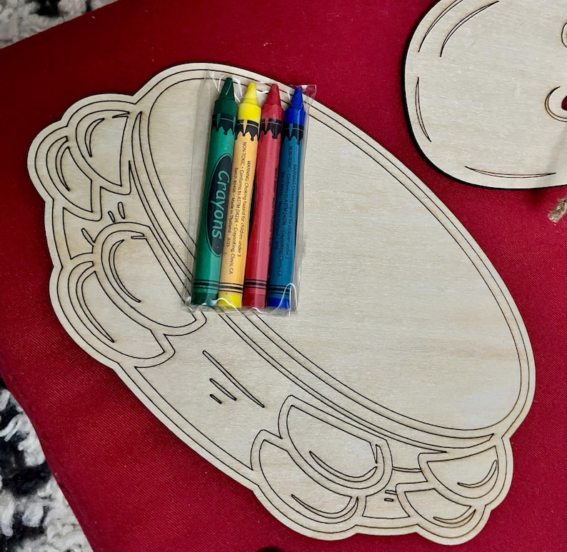 Puede incluir: Un tablero de manualidades de madera con forma de pandereta con un juego de cuatro crayones en verde, amarillo, rojo y azul envueltos en pl&aacute;stico transparente. Los crayones est&aacute;n etiquetados como "Crayons". El tablero es de madera sin terminar.