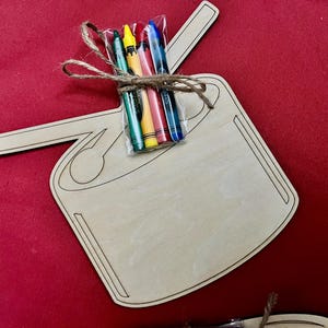 Puede incluir: Una forma de tabla de cortar de madera con un asa, atada con hilo de bramante, que contiene un paquete de crayones de colores. Los crayones son verdes, amarillos, rojos, rosas y azules. El fondo es de color rojo liso.