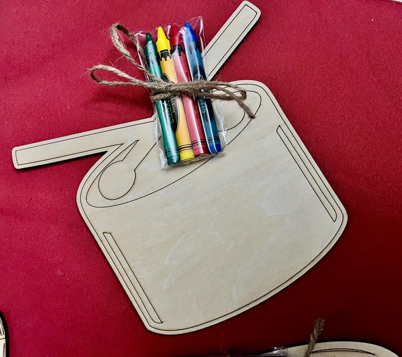 Puede incluir: Una forma de tabla de cortar de madera con un asa, atada con hilo de bramante, que contiene un paquete de crayones de colores. Los crayones son verdes, amarillos, rojos, rosas y azules. El fondo es de color rojo liso.