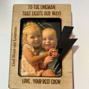 May include: A wooden picture frame with a photo of two children. The frame has the text "TO THE LINEMAN THAT LIGHTS OUR WAY!" and "LOVE, YOUR BEST CREW". A black lightning bolt is attached to the frame. The text "God Bless our Lineman" is also visible.
