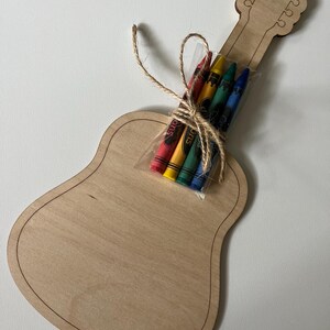 Puede incluir: Una manualidad de madera con forma de guitarra con un paquete de crayones atados con hilo. La guitarra es de madera clara con un contorno oscuro. Los crayones son rojos, amarillos, verdes y azules.