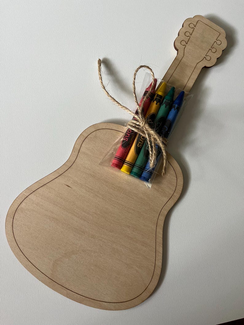 Puede incluir: Una manualidad de madera con forma de guitarra con un paquete de crayones atados con hilo. La guitarra es de madera clara con un contorno oscuro. Los crayones son rojos, amarillos, verdes y azules.