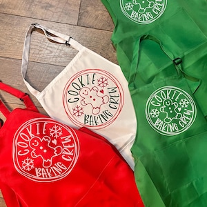 May include: Aprons in red, white, and green, each featuring a circular design with a gingerbread man and the text "COOKIE BAKING CREW." The aprons have adjustable neck straps and pockets, perfect for holiday baking.