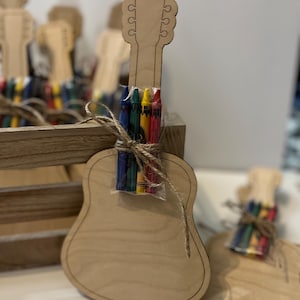 Puede incluir: Una manualidad en forma de guitarra de madera clara con un paquete de crayones de colores atados con hilo. La guitarra es de madera marr&oacute;n claro y tiene un dise&ntilde;o recortado. Se ven otras manualidades similares en el fondo, lo que sugiere un kit de manualidades o un recuerdo de fiesta.
