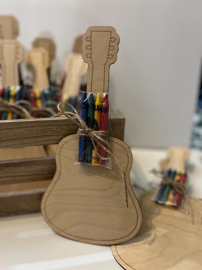 Puede incluir: Una manualidad en forma de guitarra de madera clara con un paquete de crayones de colores atados con hilo. La guitarra es de madera marr&oacute;n claro y tiene un dise&ntilde;o recortado. Se ven otras manualidades similares en el fondo, lo que sugiere un kit de manualidades o un recuerdo de fiesta.