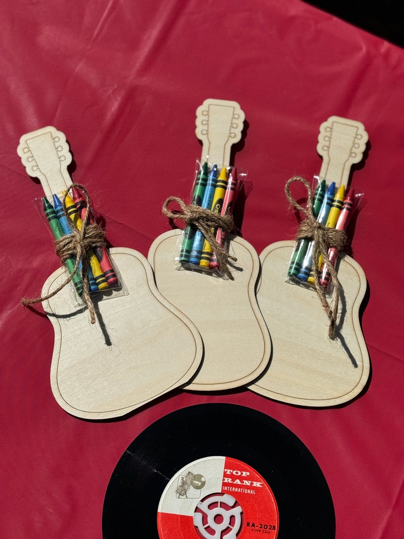 Puede incluir: Tres manualidades de madera con forma de guitarra, cada una con un paquete de crayones de colores atados con hilo. Un disco de vinilo negro con una etiqueta roja y blanca est&aacute; en la parte inferior. Las manualidades est&aacute;n sobre una superficie roja.