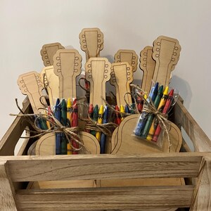Puede incluir: Una caja de madera llena de recortes en forma de guitarra y paquetes de crayones de colores. Cada paquete de crayones est&aacute; atado con hilo. Los recortes de guitarra son de color marr&oacute;n claro.