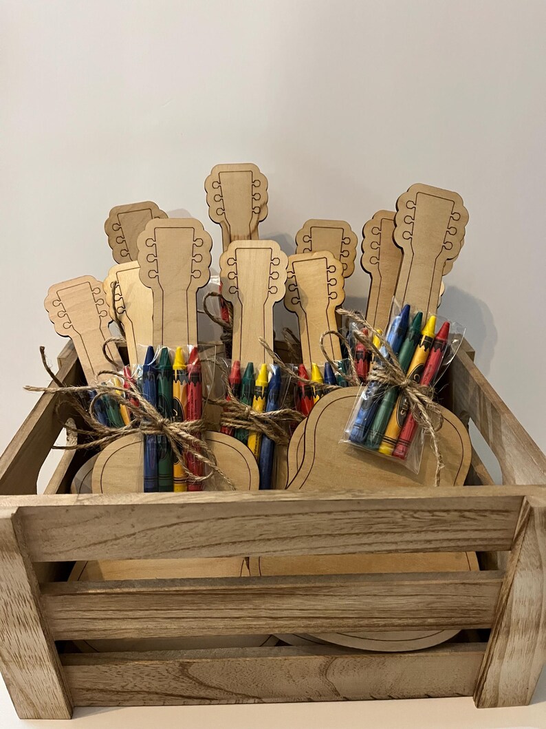 Puede incluir: Una caja de madera llena de recortes en forma de guitarra y paquetes de crayones de colores. Cada paquete de crayones est&aacute; atado con hilo. Los recortes de guitarra son de color marr&oacute;n claro.