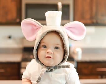 May include: A baby wearing a gray and pink mouse costume with a white chef's hat. The baby is looking at the camera and has a serious expression on their face.