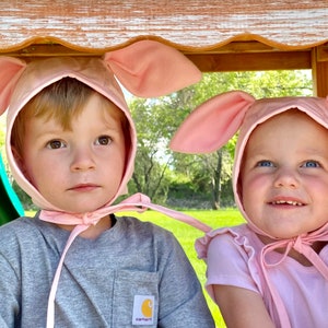 Op de afbeelding: Twee lichtroze stoffen mutsjes met grote, rechtopstaande konijnenoren bovenop. Elk mutsje heeft een ronde bovenkant, een brede rand die het gezicht omlijst en dunne stoffen linten die stevig onder de kin worden vastgestrikt.