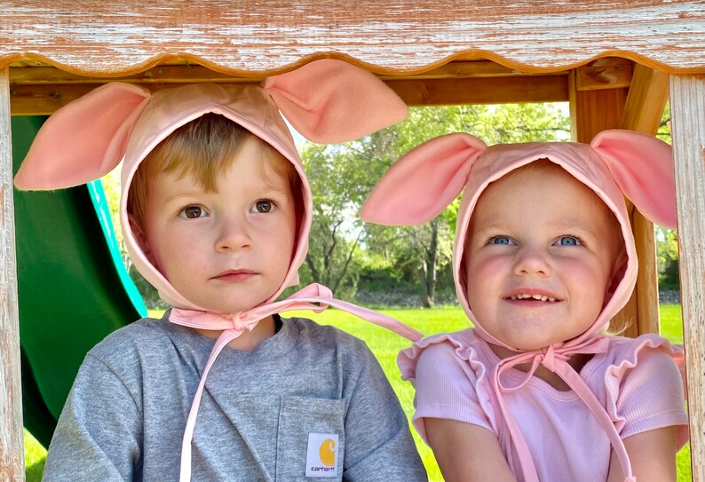 Op de afbeelding: Twee lichtroze stoffen mutsjes met grote, rechtopstaande konijnenoren bovenop. Elk mutsje heeft een ronde bovenkant, een brede rand die het gezicht omlijst en dunne stoffen linten die stevig onder de kin worden vastgestrikt.