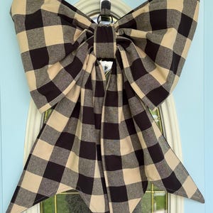 May include: A large decorative bow made of fabric with a black and beige buffalo check pattern. The bow features two wide loops at the top and two long, pointed tails hanging downwards, secured by a central fabric knot at the center point.