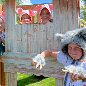 Op de afbeelding: Drie kinderen dragen lichtroze varkensmutsen met flaporen terwijl ze uit het raam van een houten speelhuisje leunen. Een vierde kind op de voorgrond draagt een grijze wolfskap van imitatiebont met bijpassende handschoenen met klauwen.