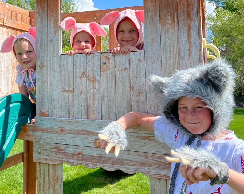 Op de afbeelding: Drie kinderen dragen lichtroze varkensmutsen met flaporen terwijl ze uit het raam van een houten speelhuisje leunen. Een vierde kind op de voorgrond draagt een grijze wolfskap van imitatiebont met bijpassende handschoenen met klauwen.
