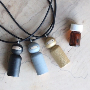 May include: Three wooden doll pendants with black, grey, and gold paint, each with a black mask and eyes, hanging on black cords. A small brown glass bottle with a white cap is in the background.