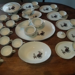 May include: A set of white porcelain teacups and saucers with a black and white design of a person riding in a rickshaw. The set includes a teapot, sugar bowl, creamer, and a large serving platter.