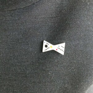 May include: White bow tie pin with black, blue, red, and yellow paint splatter design.