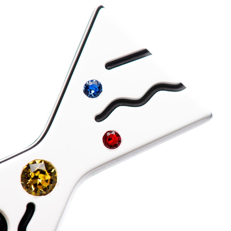 May include: A white metal bow tie with black lines and three colorful rhinestones: a blue, a red, and a yellow.