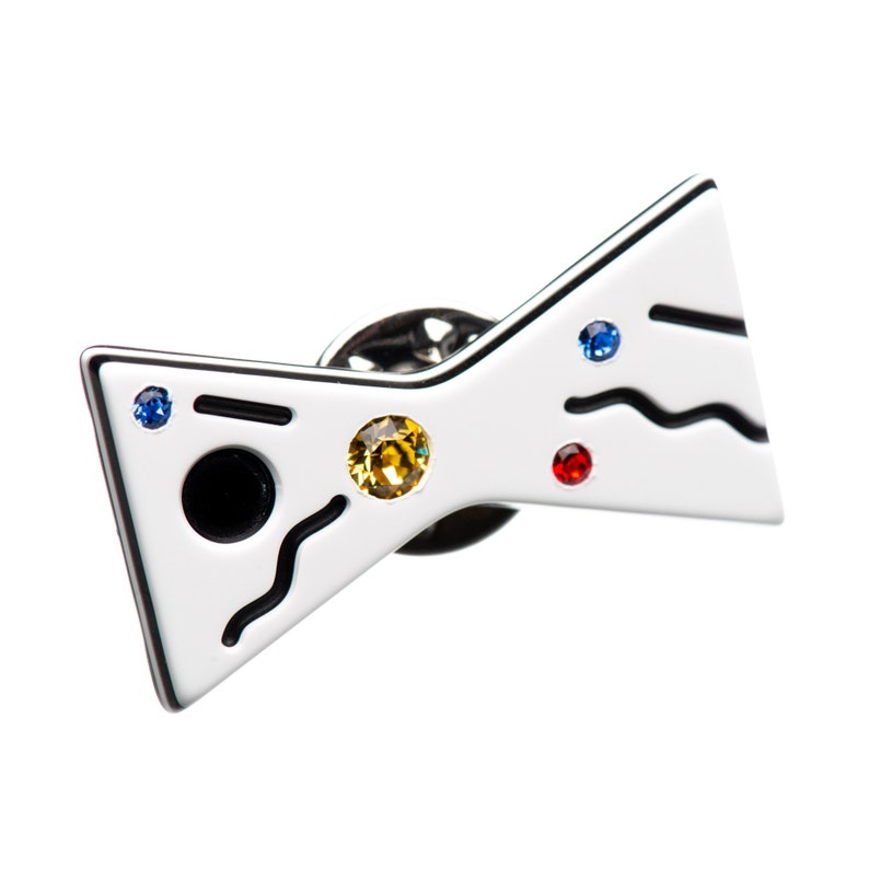 May include: White bow tie pin with black lines and colorful rhinestones. The pin has a black circle in the center.