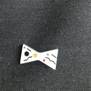 May include: A white enamel bow tie pin with black, blue, red, and yellow rhinestones.