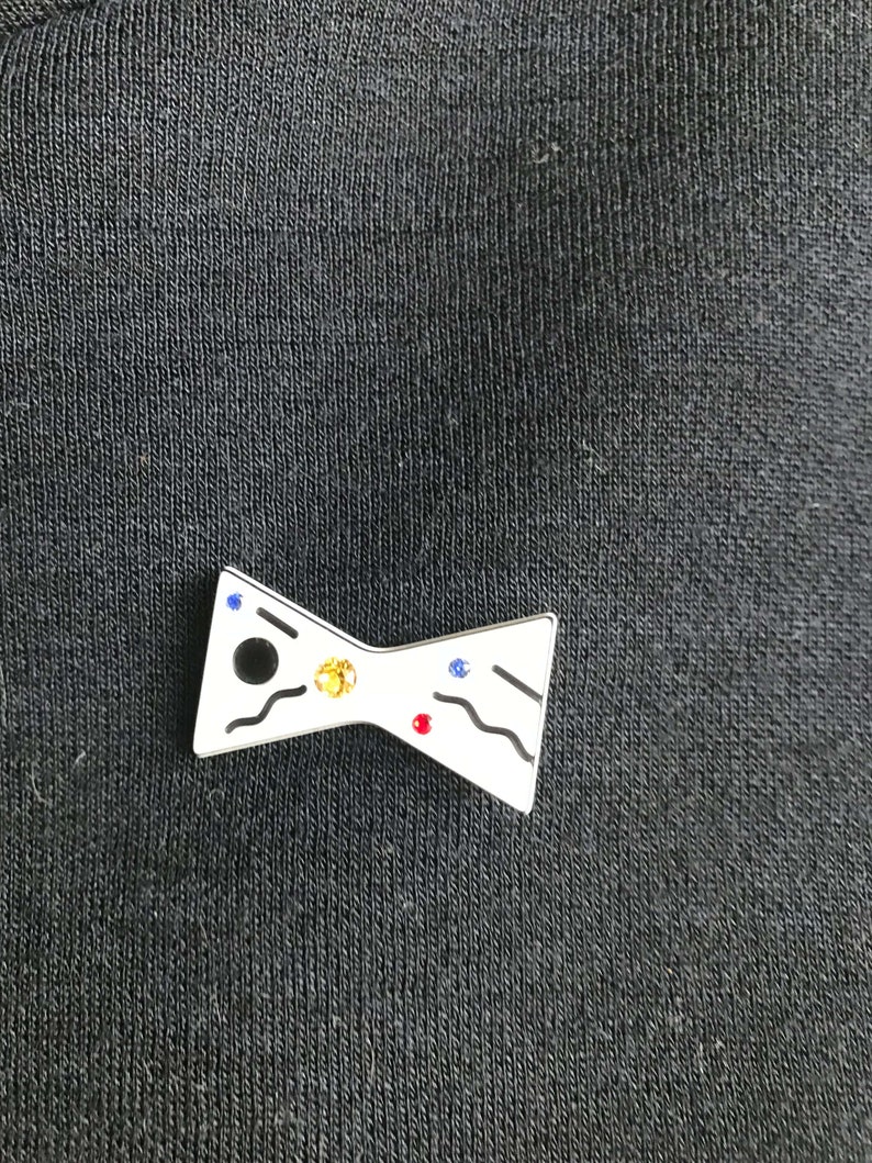 May include: A white enamel bow tie pin with black, blue, red, and yellow rhinestones.