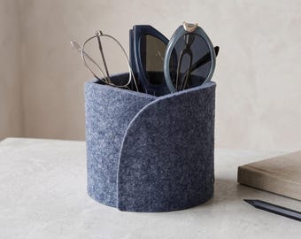 May include: A cylindrical dark gray felt desk organizer with an overlapping seam design. The container holds a pair of thin-framed metal eyeglasses and a pair of dark-tinted sunglasses with thick frames, keeping them upright and accessible.