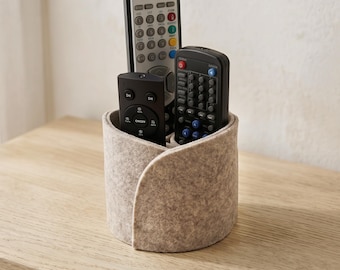 May include: A cylindrical light beige felt storage organizer holding three remote controls. The organizer features a wrap-around design with a single overlapping seam. The remote controls vary in size and color, including black and silver plastic.