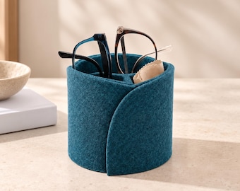 May include: A cylindrical teal felt organizer featuring a wrap-around design with internal dividers. The container holds two pairs of eyeglasses, one with blue frames and one with brown frames, along with a folded beige microfiber cleaning cloth.