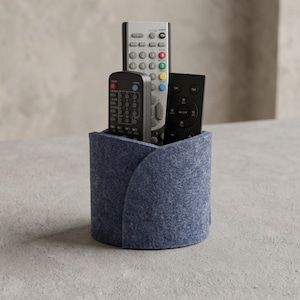 May include: A cylindrical blue felt remote control holder with an overlapping seam design. The holder contains three remote controls: one tall silver remote with multi-colored buttons, one black remote, and one smaller black remote with circular buttons.