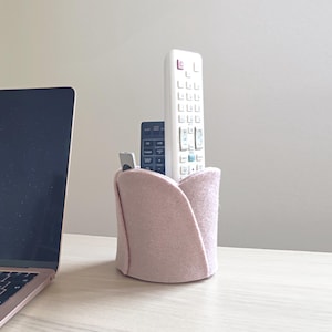 May include: A light pink felt remote control holder with a tulip-like design. Two black and white remote controls and a small silver device are inside the holder. A laptop is partially visible on the left.