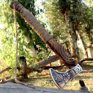 May include: A handcrafted axe with a wooden handle wrapped in twine. The axe head is silver with intricate engraved designs, including runes and a stylized wolf. A rope is attached to the handle. The axe is displayed on a log in a natural outdoor setting.