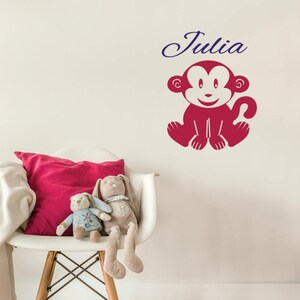 Peut inclure: Un autocollant mural de singe rouge avec un visage souriant blanc, assis sur un mur blanc avec le nom "Julia" écrit en cursive bleue au-dessus.