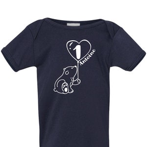 May include: A navy blue baby onesie with a white graphic of a bear holding a heart-shaped balloon with the number 1 and the name "Antoine" written on it.