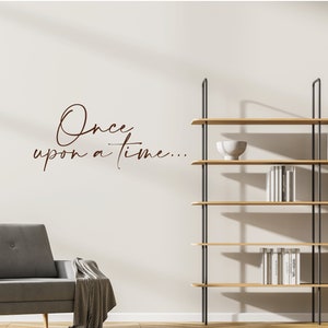 Peut inclure: Une scène de salon moderne avec un canapé gris, une étagère en bois et le texte "Once upon a time..." en écriture cursive sur le mur. L'étagère contient des livres et un bol décoratif. La pièce a une palette de couleurs neutres.