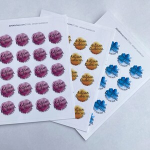 May include: Three sheets of stickers with the text "Artisan Qu&eacute;b&eacute;cois" in a watercolor-style font. The stickers are in three different color schemes: pink, orange, and blue.