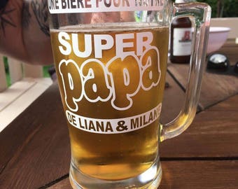 May include: A clear glass beer mug with a handle. The mug is filled with a light-colored beer. The mug has a white sticker on it that says "Une Biere Pour Notre Super Papa De Liana & Milan".