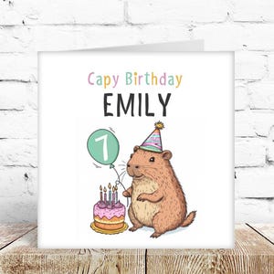 May include: A birthday card with a cartoon capybara wearing a party hat, holding a green balloon with the number 1. The card reads "Cappy Birthday EMILY" above the capybara and a birthday cake with candles. The card is on a wooden surface.