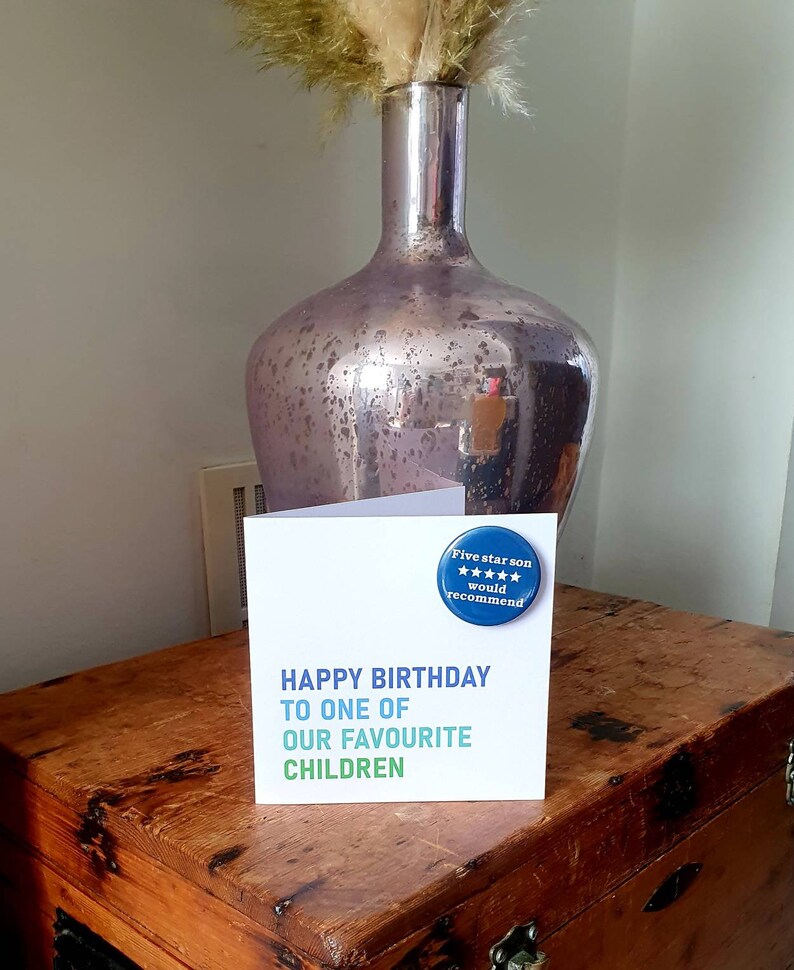 Op de afbeelding: Een witte kaart met de tekst "Happy Birthday to one of our favourite children" en een blauwe knop met de tekst "Five star son would recommend" en vijf sterren.