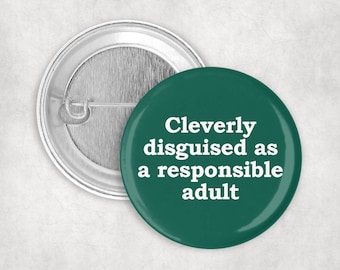 May include: A round pinback button with a dark green front featuring the white text "Cleverly disguised as a responsible adult" in a serif font. The back of the button is metallic silver with a standard pin mechanism for attachment.