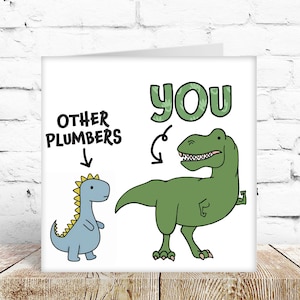 May include: A white greeting card with a dinosaur theme. The card features two cartoon dinosaurs, one small and blue labelled "OTHER PLUMBERS" and a large green dinosaur labelled "YOU". The word "YOU" is in green with a textured design.