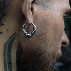 Puede incluir: Un hombre con la cabeza rapada y barba tiene tatuajes geométricos en la cabeza y el cuello. Lleva un aro de plata en la oreja izquierda.