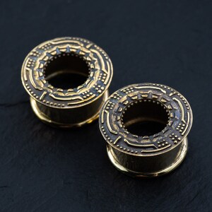 May include: Two gold-colored metal ear gauges with intricate geometric designs. The gauges are on a black surface.