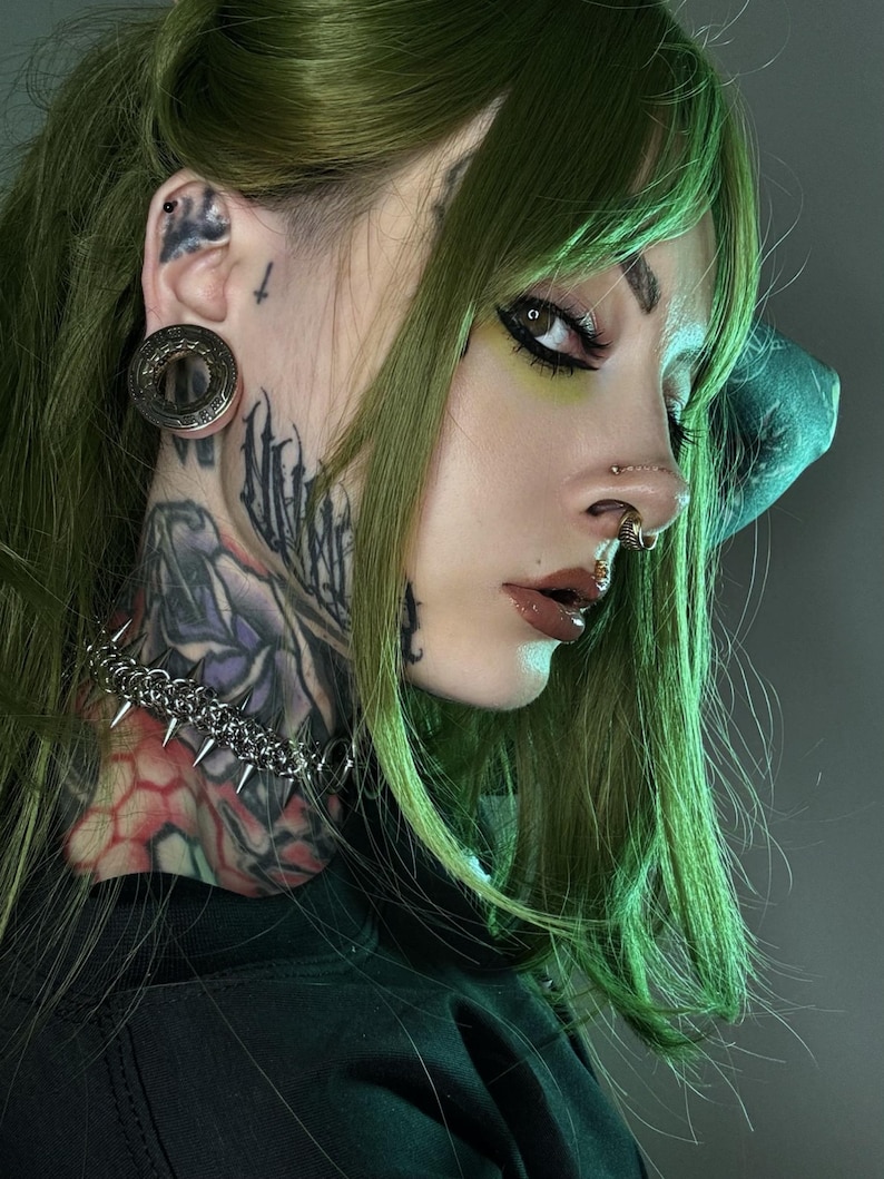 A woman with green hair and multiple facial and ear piercings, including a large gold tunnel with an alien spaceship design. She has heavy neck tattoos, a spiked chain choker, and a cyberpunk aesthetic with futuristic, HR Giger-inspired style.