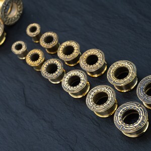 May include: A set of gold-colored ear gauges with intricate designs. The gauges are arranged in a row from smallest to largest, with the smallest gauge at the top of the image and the largest gauge at the bottom.