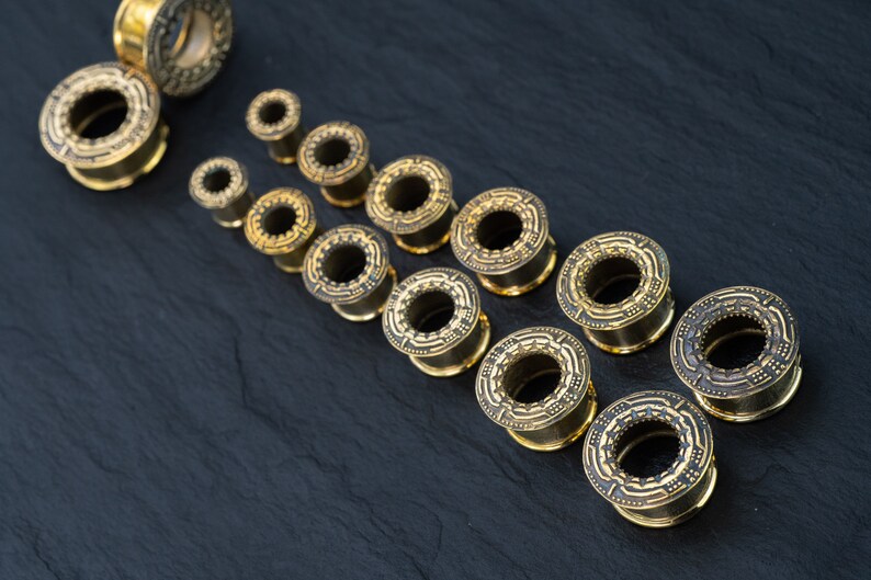 May include: A set of gold-colored ear gauges with intricate designs. The gauges are arranged in a row from smallest to largest, with the smallest gauge at the top of the image and the largest gauge at the bottom.