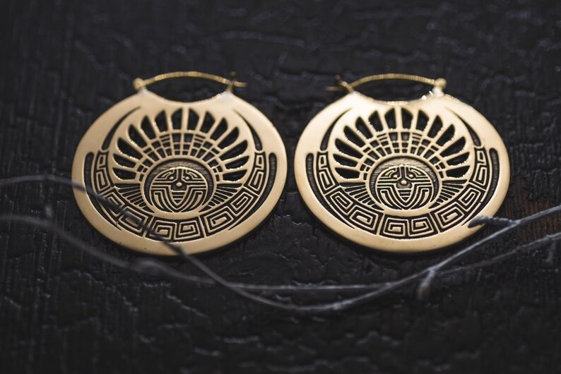 Puede incluir: Un par de grandes pendientes de aro dorados con un dise&ntilde;o geom&eacute;trico. Los pendientes presentan un patr&oacute;n circular con un dise&ntilde;o de sol en el centro.
