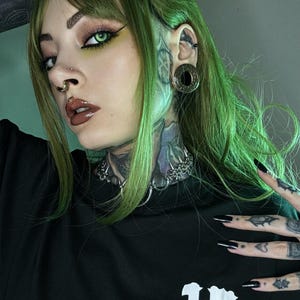 A person with green hair, a nose ring, and a large gold tunnel in their stretched ear. They have tattoos on their neck and hands. Their look combines bold, futuristic, and cyberpunk elements, with sharp, detailed styling