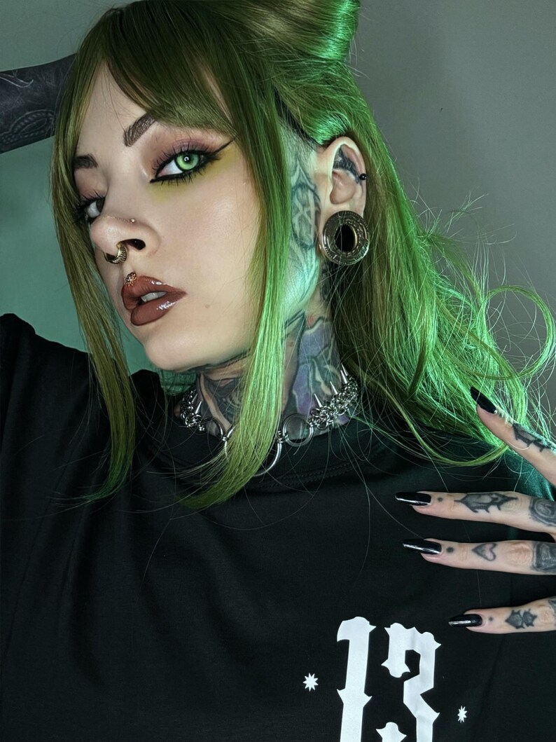 A person with green hair, a nose ring, and a large gold tunnel in their stretched ear. They have tattoos on their neck and hands. Their look combines bold, futuristic, and cyberpunk elements, with sharp, detailed styling