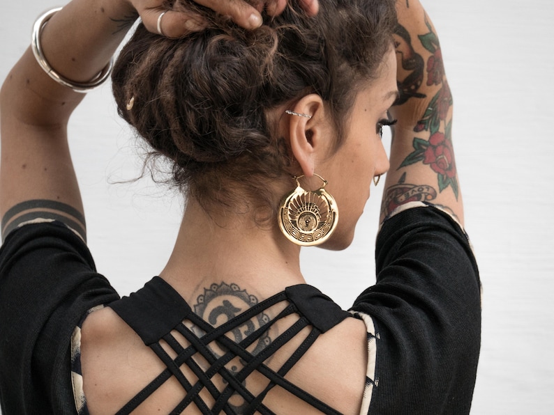 Puede incluir: Una mujer con tatuajes y un mo&ntilde;o en el pelo lleva una blusa negra con un dise&ntilde;o de espalda cruzada. Lleva grandes pendientes de aro dorados con un dise&ntilde;o circular.